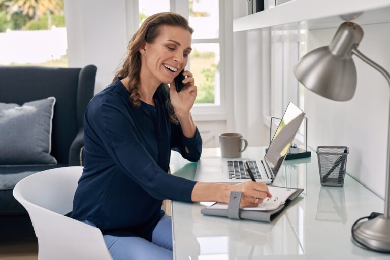 Casual businesswoman working remotely from home office writing on notepad and talking on mobile phone.