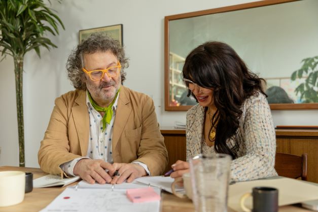 Two people collaborate at a table, reviewing documents and notes with smiles, in a bright, inviting workspace adorned with greenery.