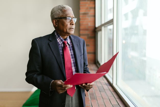 An older man in a suit and red tie stands by a window, holding red folders and gazing thoughtfully outside. The image suggests a moment of contemplation or decision-making.