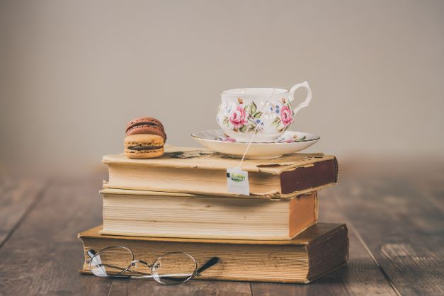Teacup with floral design sits on a stack of vintage books, accompanied by a macaroon and reading glasses. A cozy scene for book lovers.